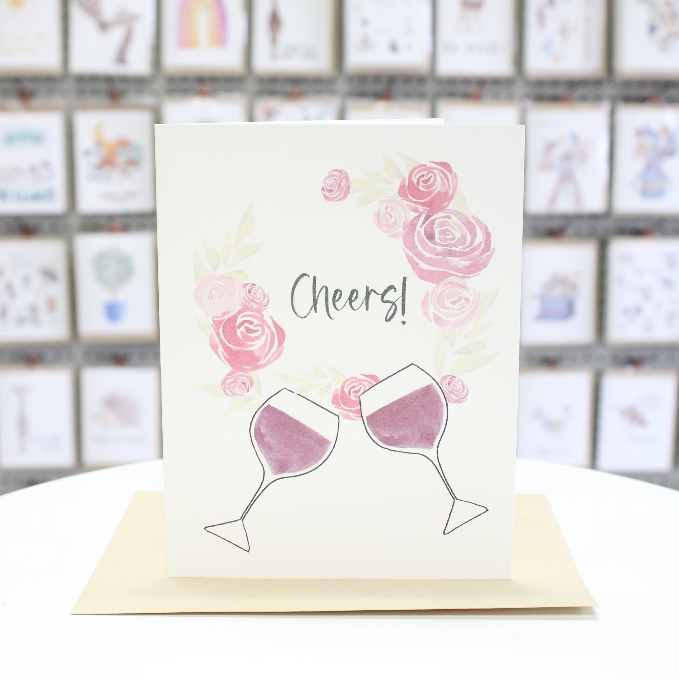Hand crafted greeting card with a wreath of watercolor roses and two wine glasses. The words, "Cheers" inside the wreath. 