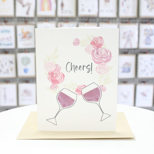 Hand crafted greeting card with a wreath of watercolor roses and two wine glasses. The words, "Cheers" inside the wreath. 