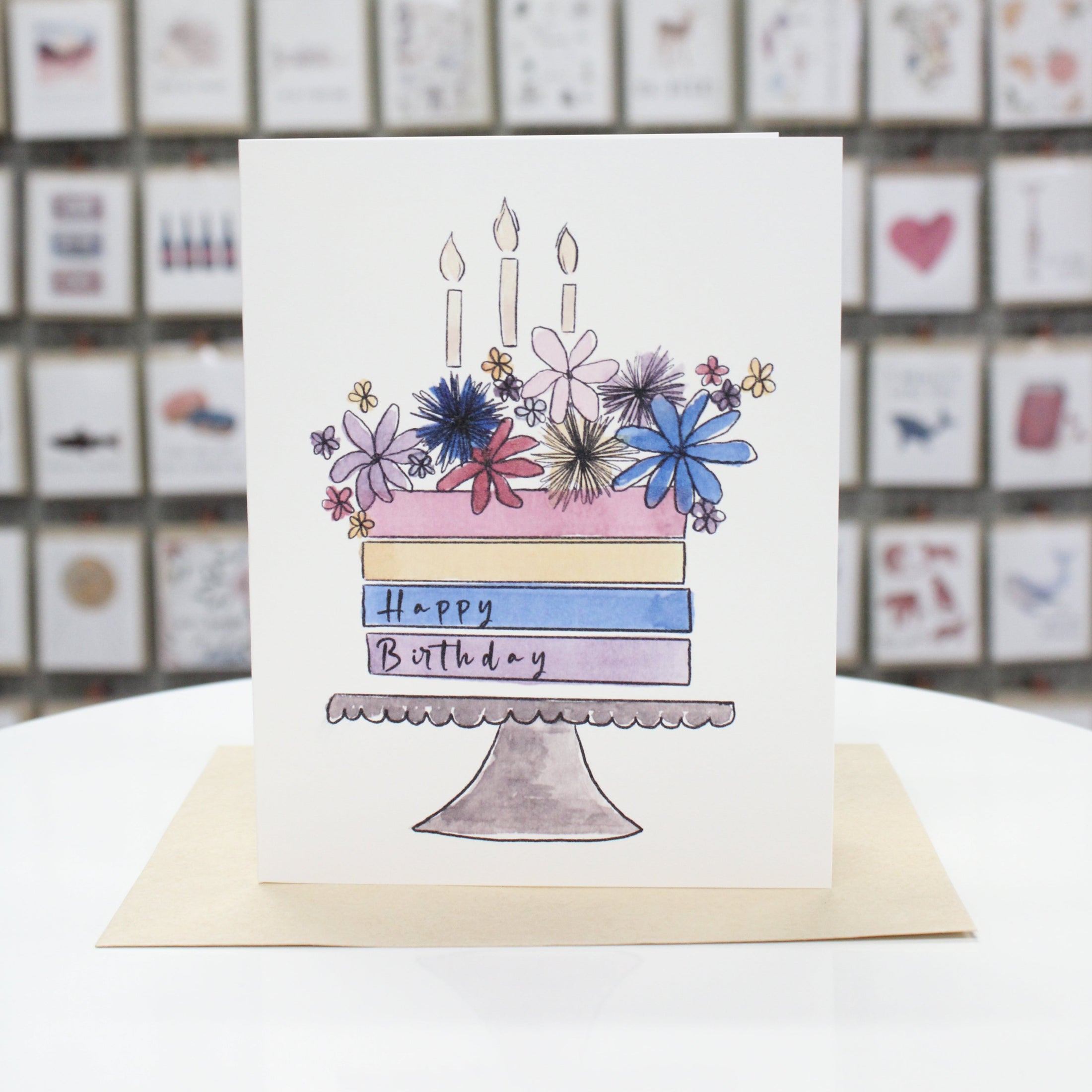 Hand crafted birthday card. Hand drawn and water colored birthday cake on a display plate with candles and flowers on the top. On the cake states, "Happy Birthday."